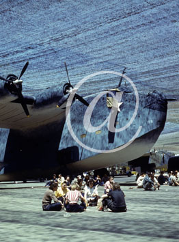 inconnu() Seconde guerre mondiale US Navy en couleurs - Srie 80-GK- : Ouvriers au repos assis sur une piste devant un gros porteur camoufl par un filet tendu au dessus de la piste.